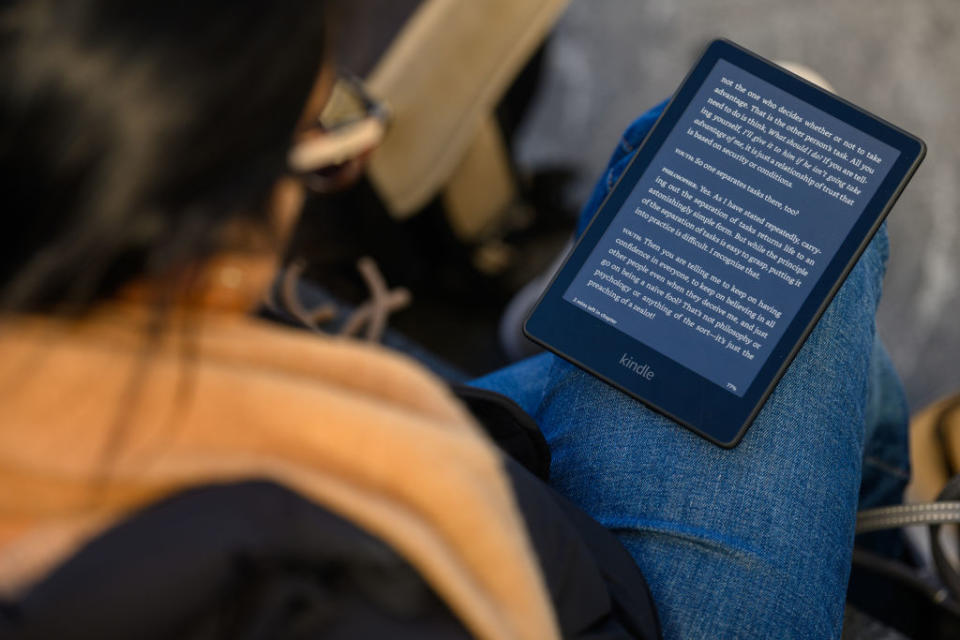 A person, viewed from above, reads an Amazon Kindle device with a full page of text. The person wears glasses and a scarf