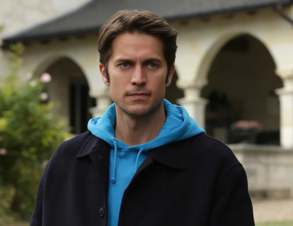 Lucas Bravo, dressed in a casual jacket and hoodie, stands in front of an arched building with greenery in the background in "Emily in Paris"