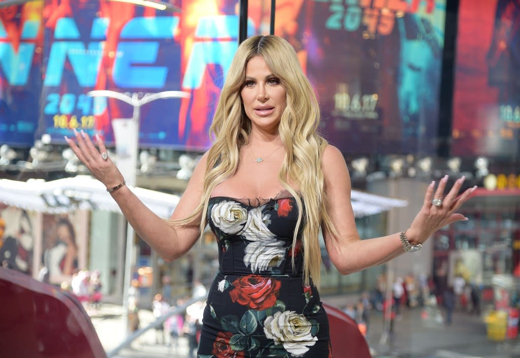 Kim Zolciak on Extra, wearing a floral dress and holding her hands up, shrugging her shoulders