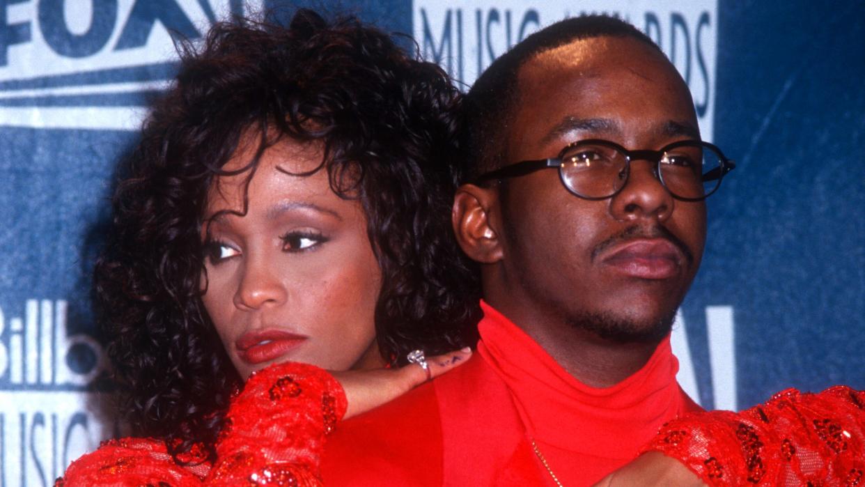 whitney houston and bobby brown pose for a photo, she hugs him from behind with her arms on his shoulders and chest, they wear matching bright red outfits