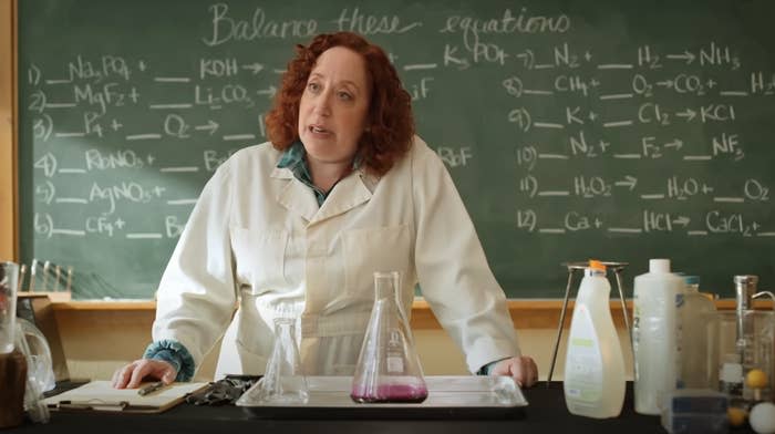 Woman in a lab coat stands behind a lab table with glassware and chemicals. A chalkboard filled with chemical equations is behind her