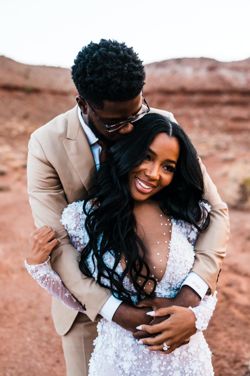 A groom hugs his bride from behind and kisses her forehead while she smiles.