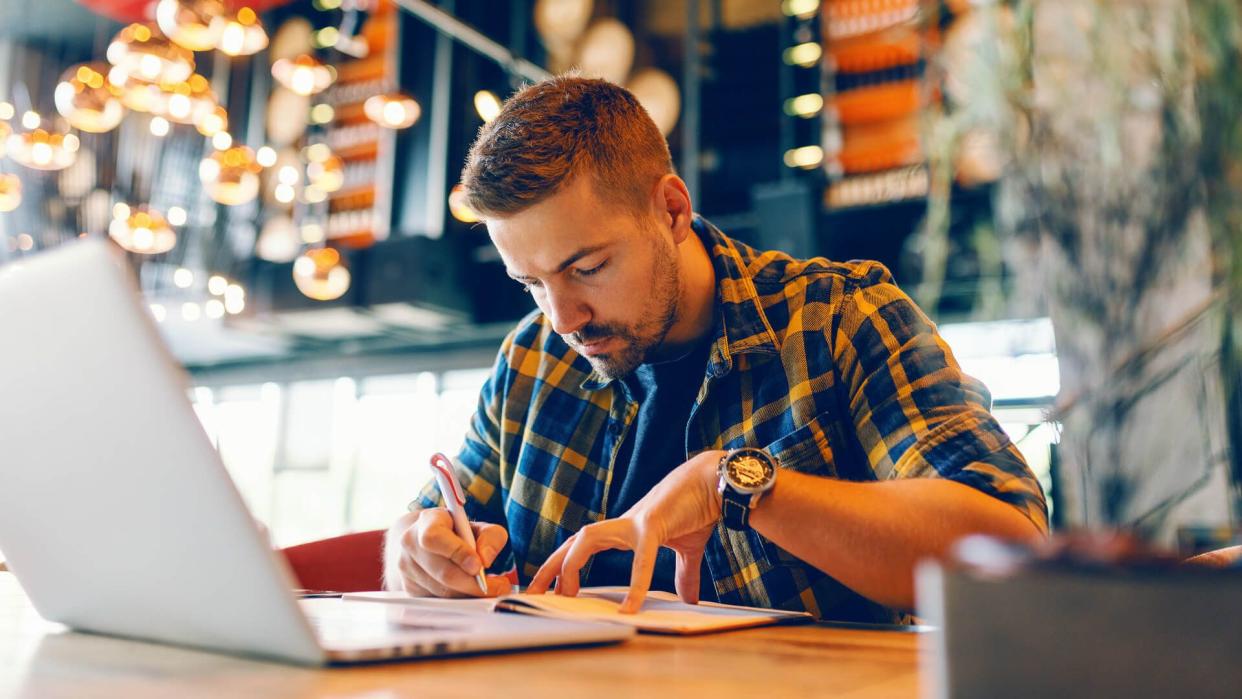 Focused bearded Caucasian freelancer writing in notebook tasks while sitting in cafeteria.