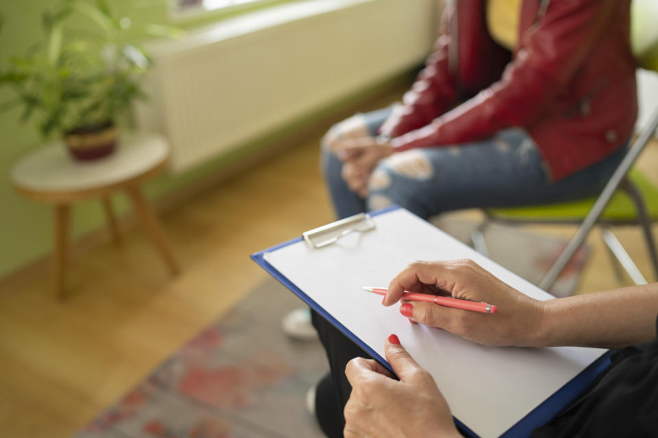 Two people sitting in a room; one person is holding a clipboard and a pen, appearing to take notes while the other is in casual attire