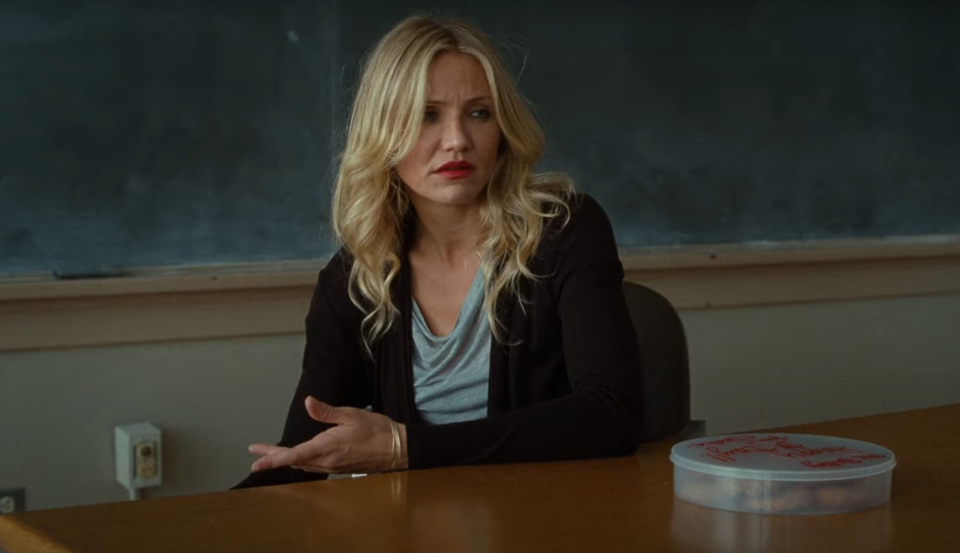 Cameron Diaz sitting at a classroom desk, gesturing with one hand, with a container of cookies on the desk in front of her