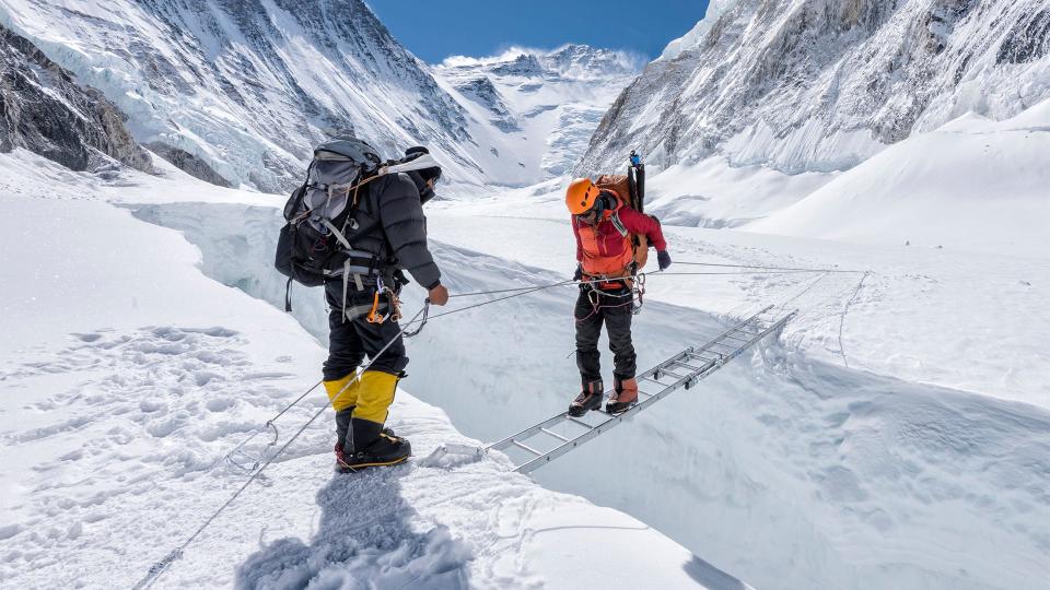 Two mountain climbers cross an icy crevice using a horizontal ladder.