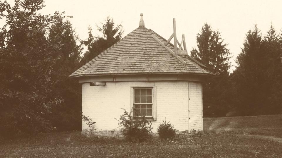 A sepia-toned historical photo of a small hut in a grass field surrounded by pine trees.