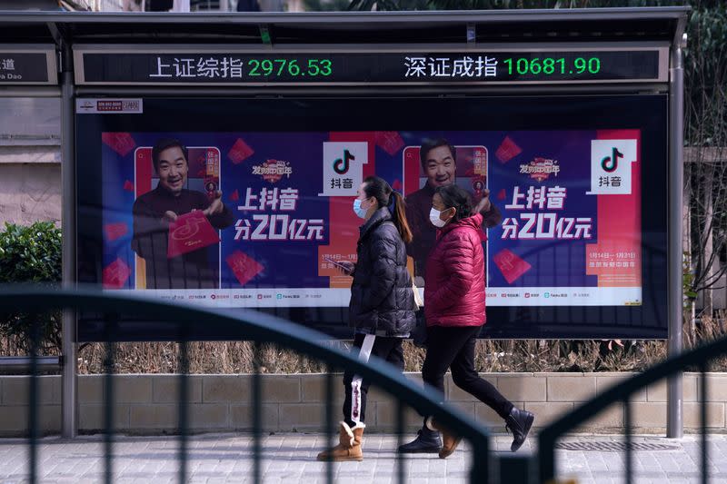 Women wearing masks walk in front of an electronic board showing the Shanghai and Shenzhen stock indexes in Shanghai