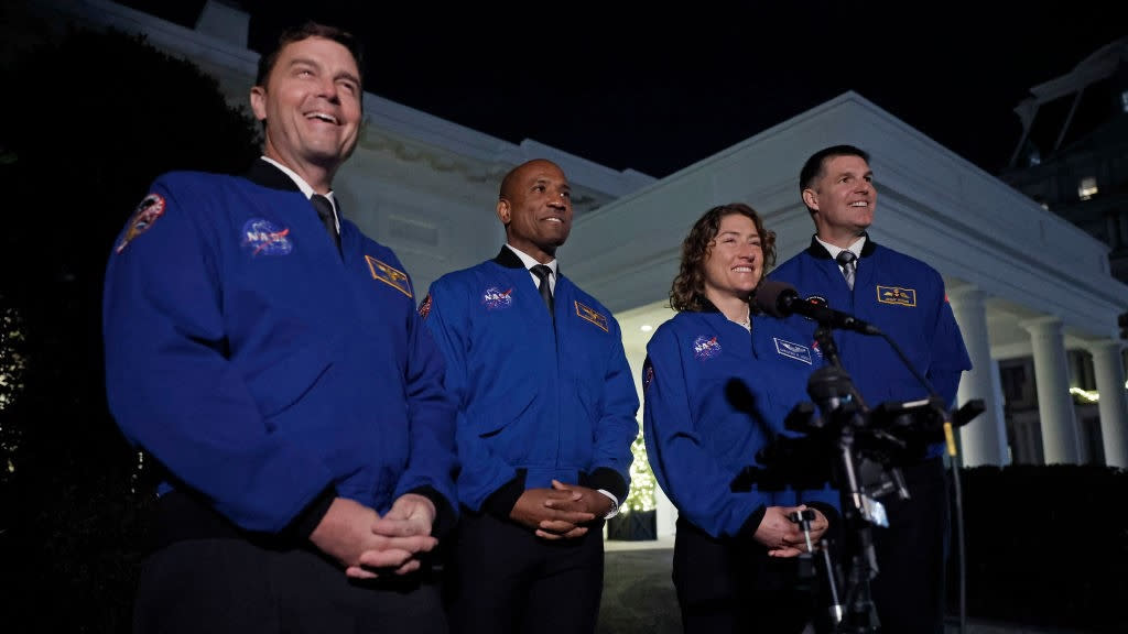  Four humans in blue jackets smile before microphones, in front of a white building. . 