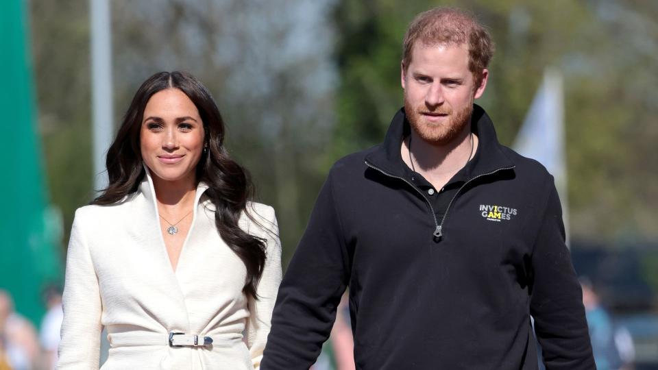 Meghan Markle in a white coat and jeans walking with Prince Harry in a black jacket and tan trousers