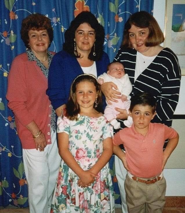 A family photo with three adults, one holding a baby, and two children in the front, a girl smiling and a boy looking annoyed, with his hands on his waist