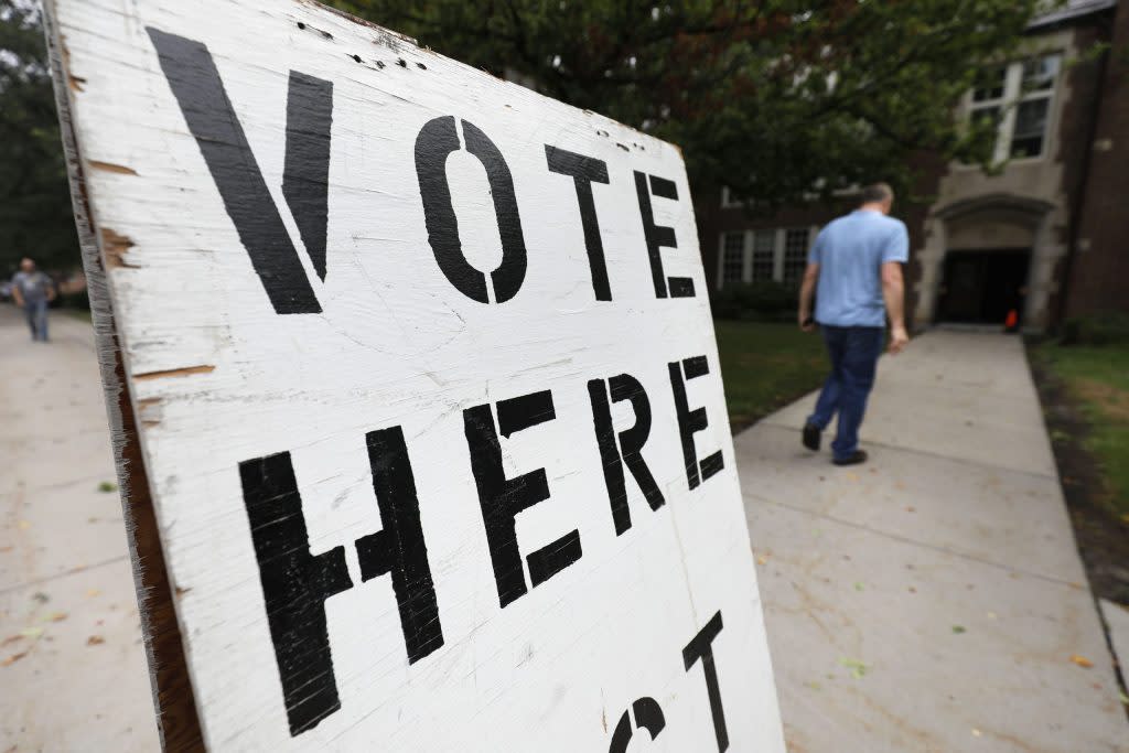 A sign saying "Vote Here" in black lettering on a white board.