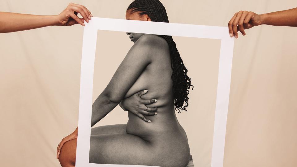Naked woman sitting sideways behind a picture frame. Confident young woman embracing her natural body and curves against a studio background. Two hands holding up a white picture frame in a studio.