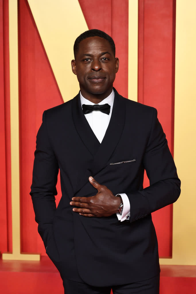 Man in a tailored black tuxedo with bow tie, standing, hand over chest, at an event