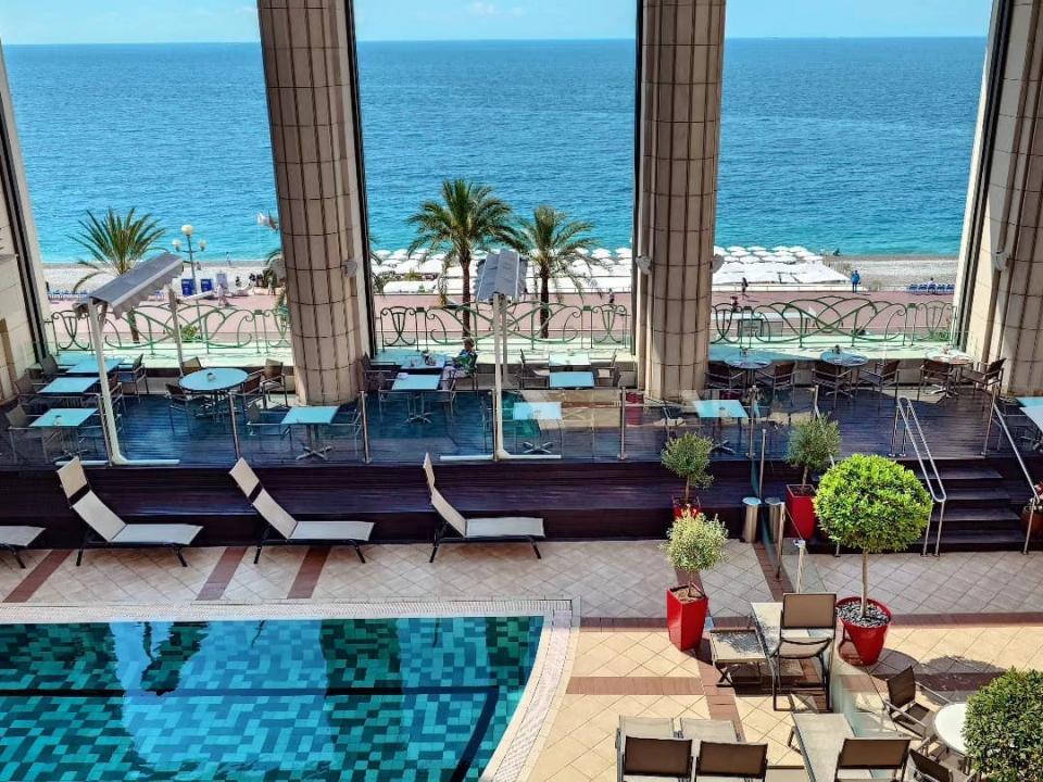Ein Hotel mit einem Pool, mehreren Liegestühlen, Topfpflanzen und großen Fenstern mit Blick auf die französische Riviera. Das Wasser ist leuchtend blau