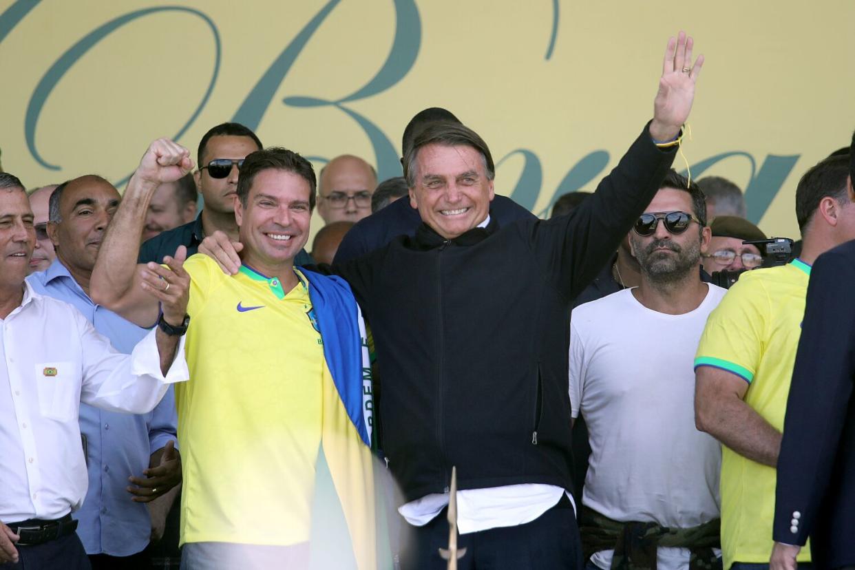 A man in a dark jacket, right, smiles next to another man in a yellow-and-green shirt as they are surrounded by others