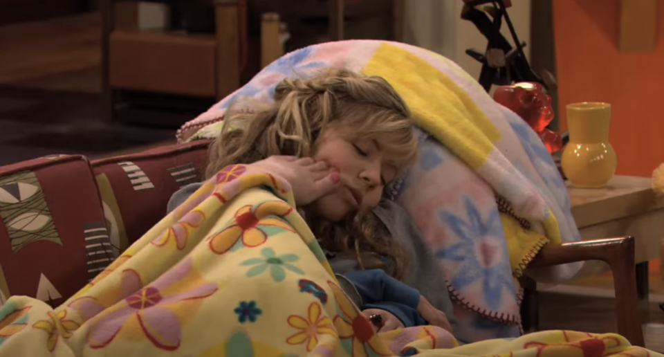 Child character resting on a couch under a blanket in a sitcom scene