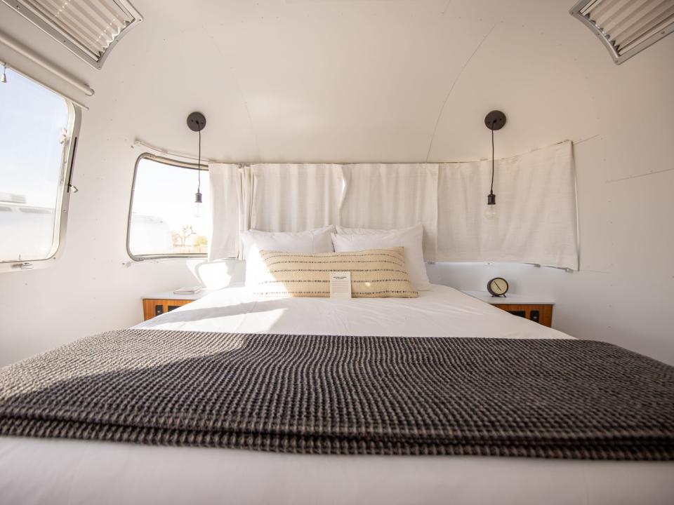 A bedroom inside Autocamp Joshua Tree's Airsteram trailer with white accents, a bed with pillows, curtains over windows.