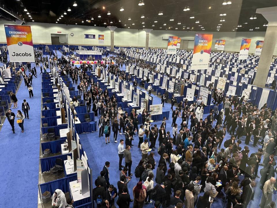 high school students in business clothes crowd and line up at one side of a giant exhibit hall with blue carpet filled with lines of poster boards