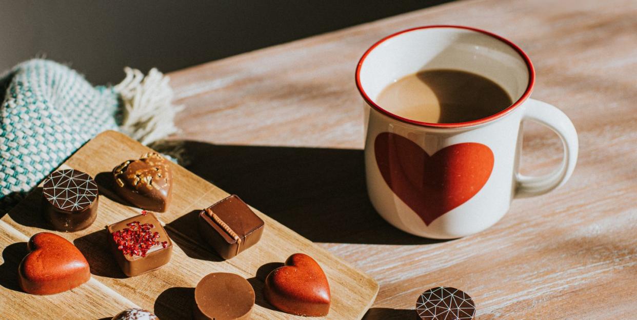 luxury chocolates in various shapes and flavours, including red heart chocolates they sit on wooden surfaces, beside a luxurious wool blanket beside the sweets sits a hot mug of coffee with a large heart emblem