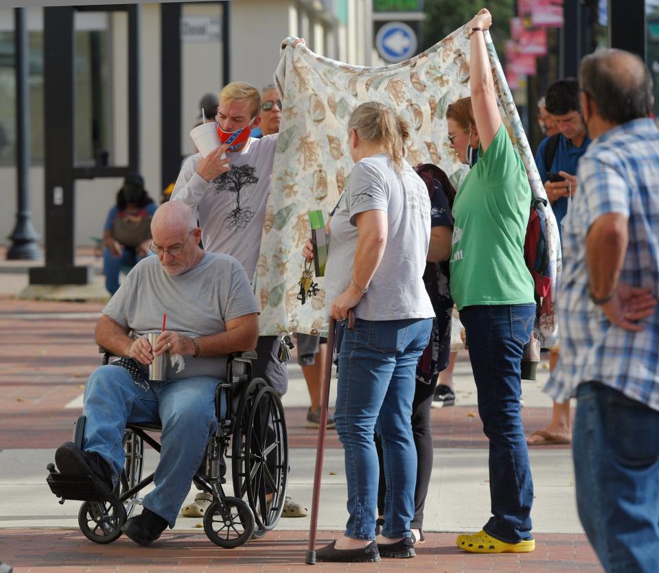 Family members provide shade with a sheet for a loved one as they waited on the sidewalk outside the main library for the Regeneron clinic inside to open Friday morning. About 50 people were in line outside the downtown Jacksonville, Florida main library Friday, August 20, 2021 as they waited for the 9 am opening of the  clinic inside that gives Regeneron's Monoclonal Antibody treatment to mitigate the symptoms of COVID-19. [Bob Self/Florida Times-Union]