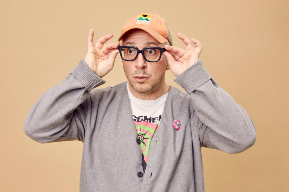 Steve adjusts his eyeglasses as he looks off-camera. He's wearing a baseball cap, a band shirt, and a sweater