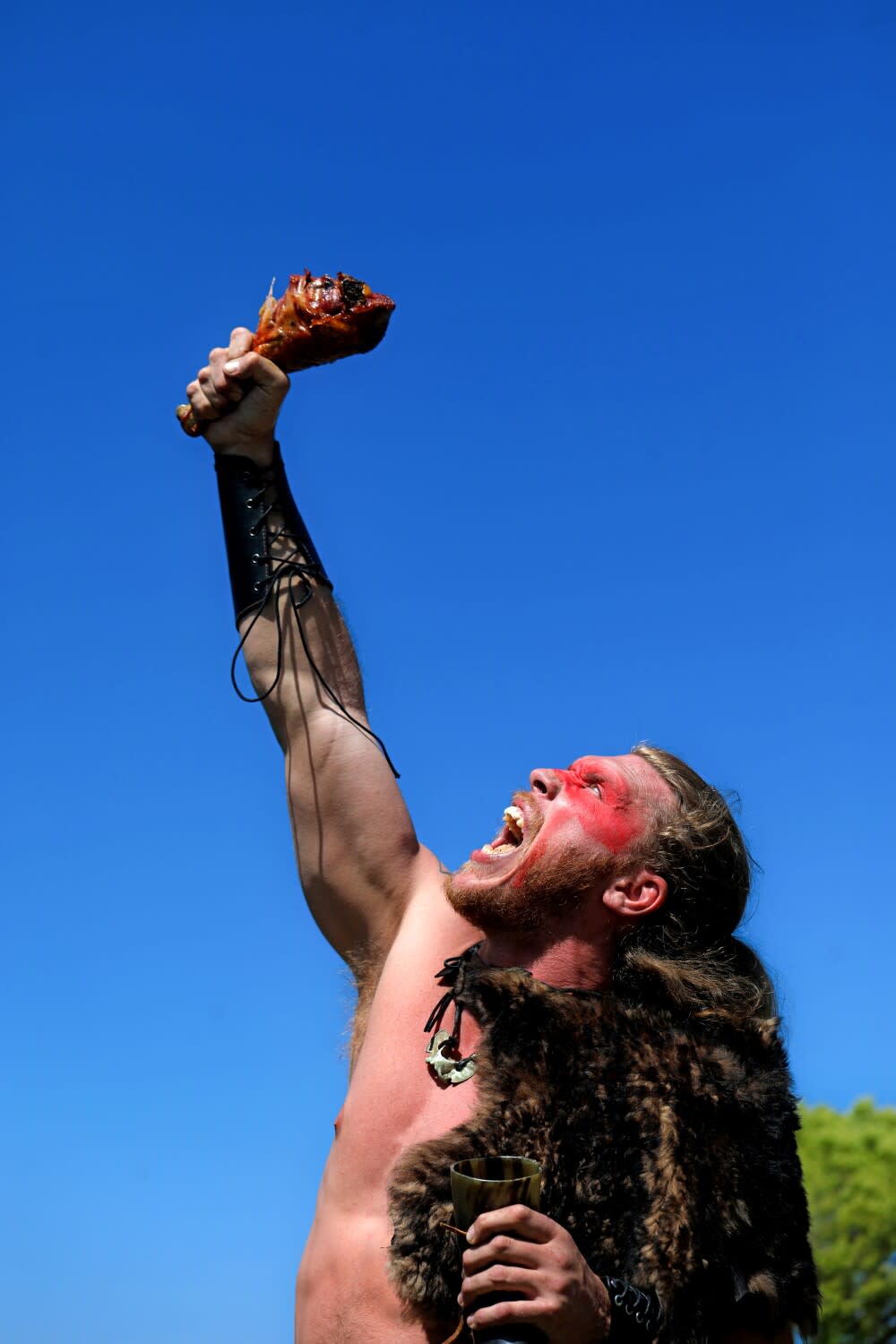 A man in an animal-pelt cape holds a turkey leg above his head