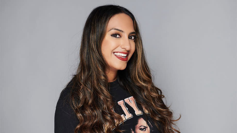 Jasmine Takhar, an Asian woman, smiling. She is wearing a black jumper with pink writing partially visible and partly covered by her hair. The background is plain grey.