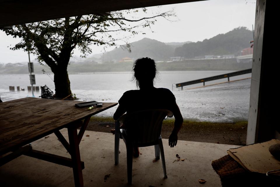 A silhouette of a man sitting in a chair looking out towards a body of water, looking at the heavy rain falling.