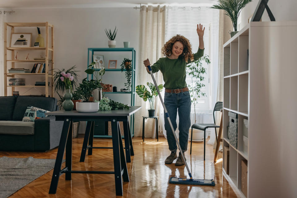 Woman with curly hair mopping the floor, smiling and looking energized, in a stylish, plant-filled living room