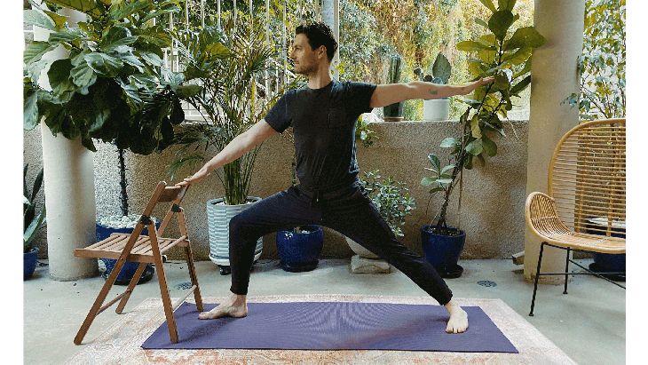Man standing on a yoga mat with a chair in front of him practicing Warrior 2 Pose with the chair as added support for balance