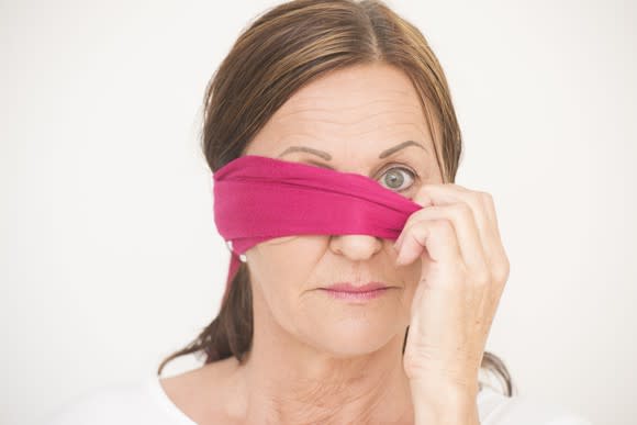 mature woman peaks out one eye from behind a purple blindfold.