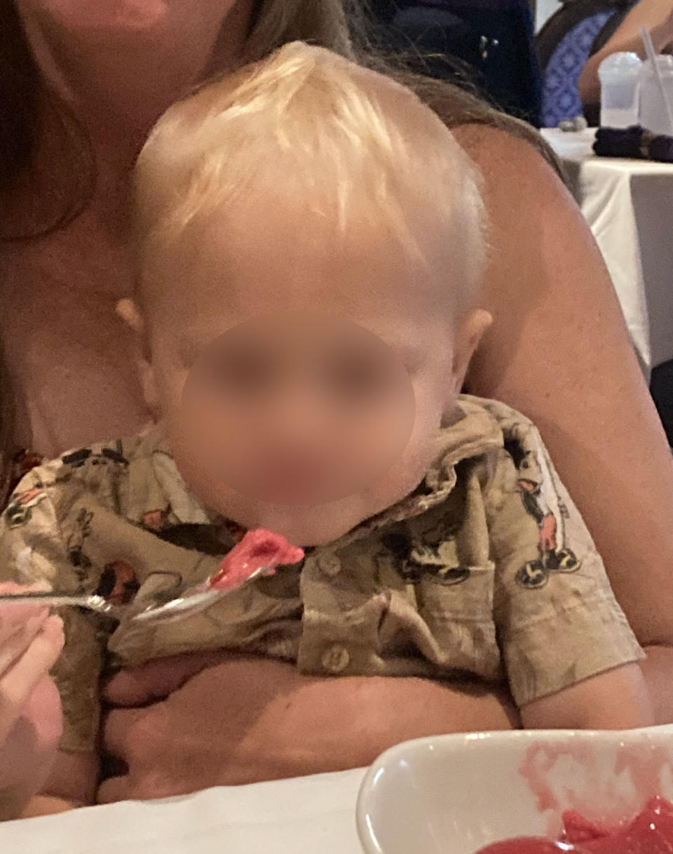 A baby wearing a patterned shirt is being fed with a spoon by an adult. The baby appears to be eating a pink substance