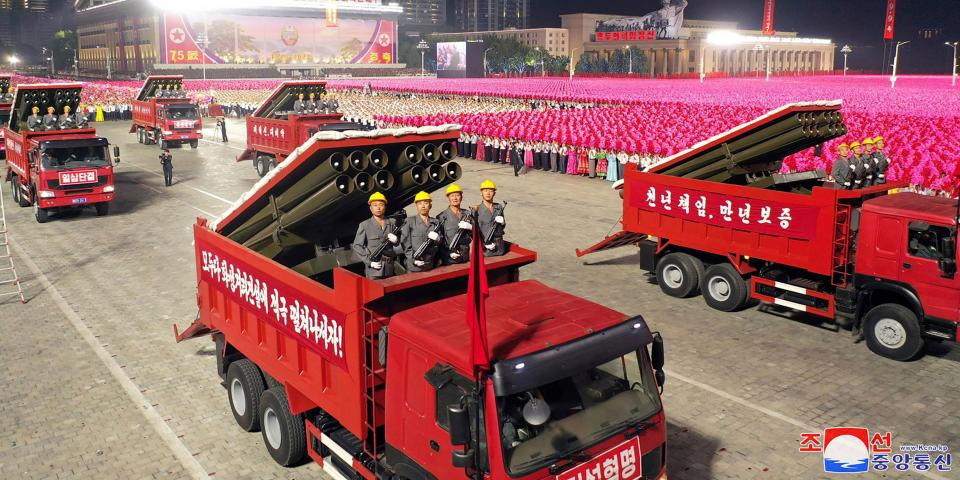 red truck in foreground features four people in gray suits and yellow helmets emerging from back, alongside missile launchers. more such trucks surround it, with crowds in match pink dress standing in distance