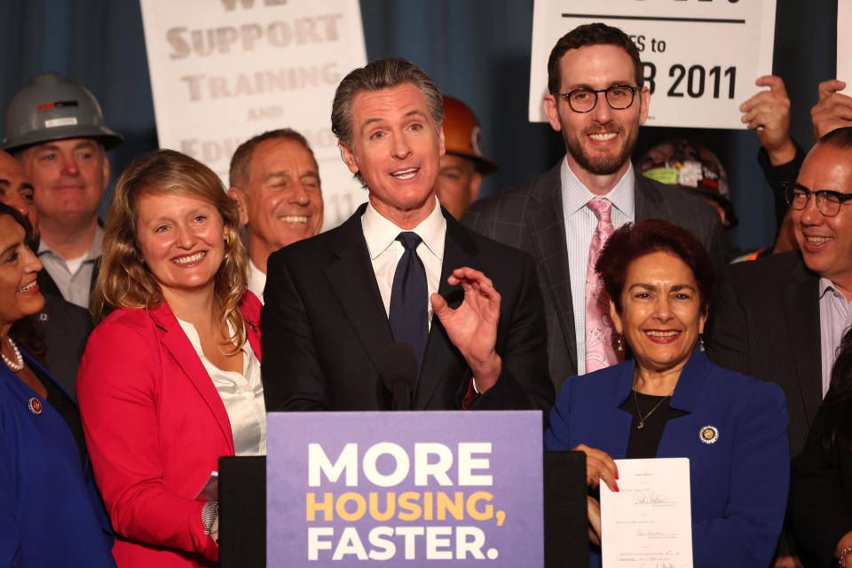 California Gov. Gavin Newsom surrounded by supporters in front of a dais saying: More Housing, Faster.