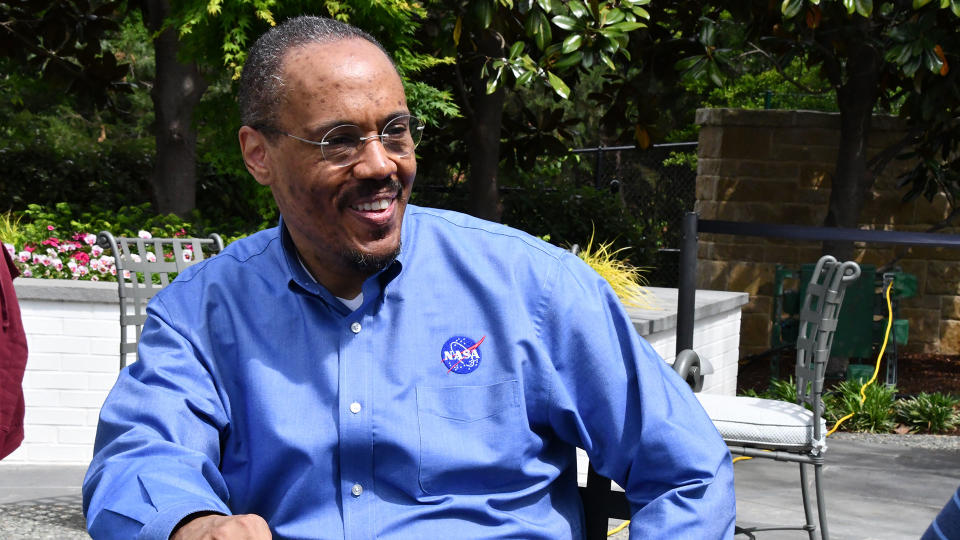 a smiling man in a blue collared shirt sits in a chair