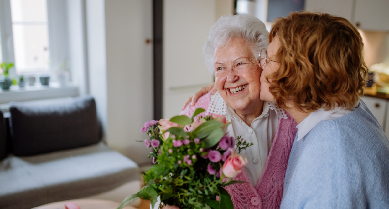 Mother's Day, woman with short red hair and blue sweater kissing elderly woman on cheek with white hair holding bouquet of flowers, mother's day flowers 