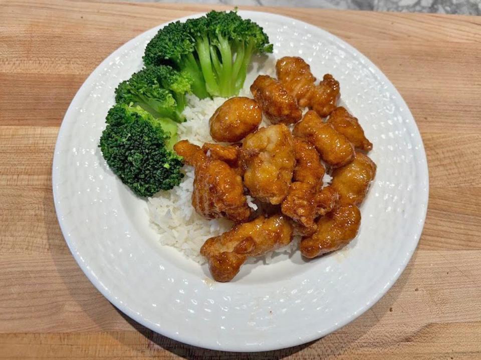 A white plate with white rice, broccoli, and pieces of orange chicken placed on a wooden cutting board