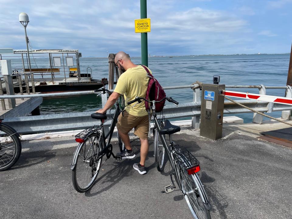 A man wearing a yellow shirt and red backpack in between two bikes on sea port in Italy