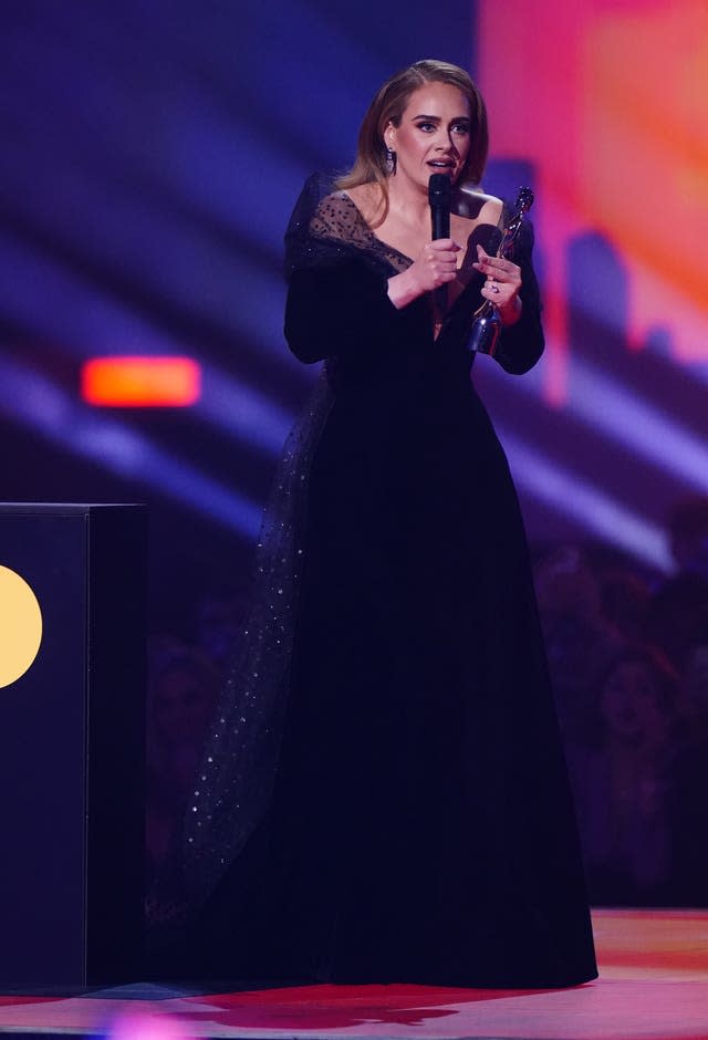 Adele on stage wearing a black dress and holding a microphone and statuette