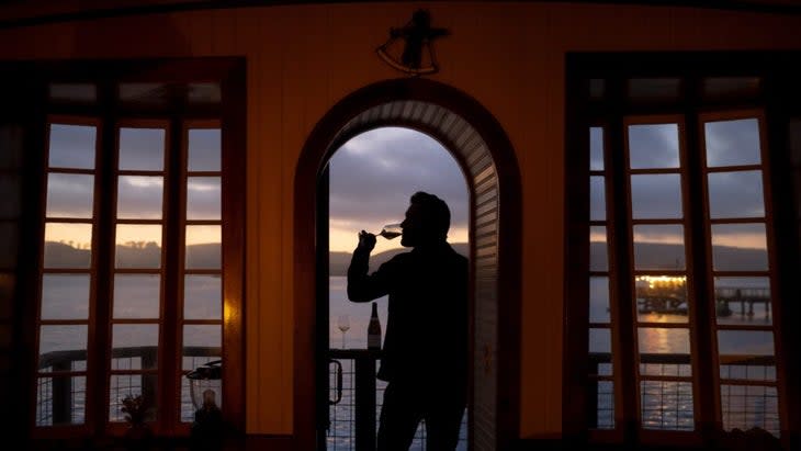 A man drinking a glass of wine in the doorway of one of the cottages, with Tamales Bay and the boat shack in the background