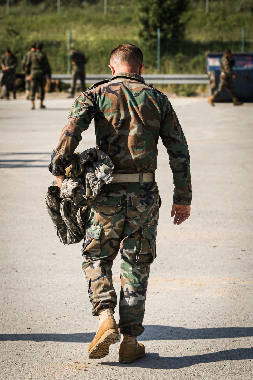 An image of a man in army fatigues walking with his head down and holding fire-resistant clothing in his left hand.