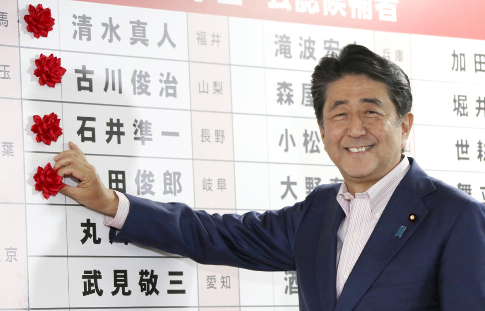 Japanese Prime Minister Shinzo Abe smiles in front of red rosettes on the names of his Liberal Democratic Party's winning candidates during ballot counting for the upper house elections at the party headquarters in Tokyo, Sunday, July 21, 2019. Prime Minister Abe's ruling coalition appeared certain to hold onto a majority in Japan's upper house of parliament, with exit polls from Sunday's election indicating he could even close in on the super-majority needed to propose constitutional revisions.(AP Photo/Koji Sasahara)