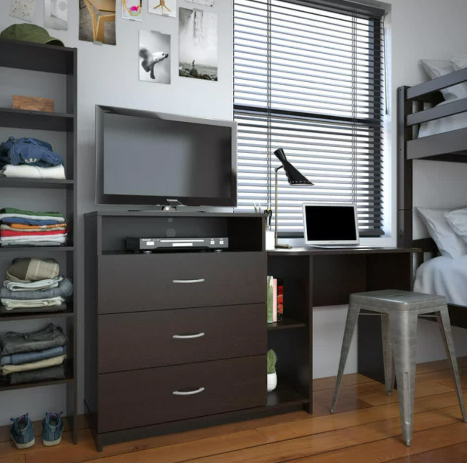 A tidy, modern bedroom with a TV, computer on a desk, window with blinds, and organized shelves