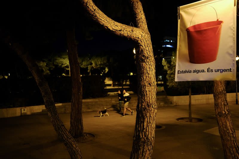 Un grupo de personas pasa corriendo con su perro junto a un cartel en el que se lee "Ahorra agua, es urgente", alertando de la grave sequía en la región noreste de España e instando a ahorrar agua debido a los bajos niveles de los embalses, en un momento en que Cataluña declara el estado de emergencia, en Barcelona, España