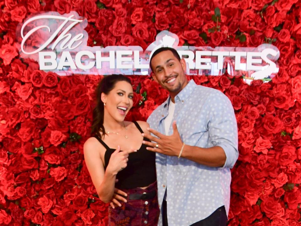 Becca Kufrin and Thomas Jacobs post in front of a wall of red roses