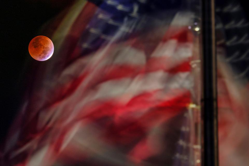 The full blood moon caused by the lunar eclipse is framed by the US Flags blowing in the breeze on the National Mall in Washington, Tuesday, Nov. 8, 2022.