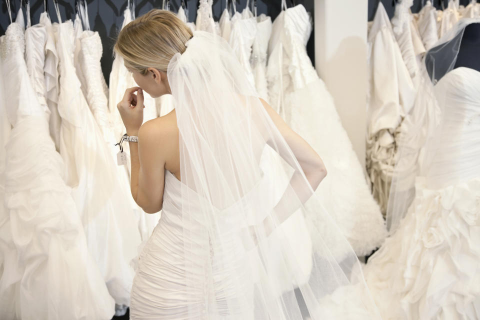 A bride in a wedding dress with a long veil stands with her back to the camera, surrounded by other wedding dresses on hangers