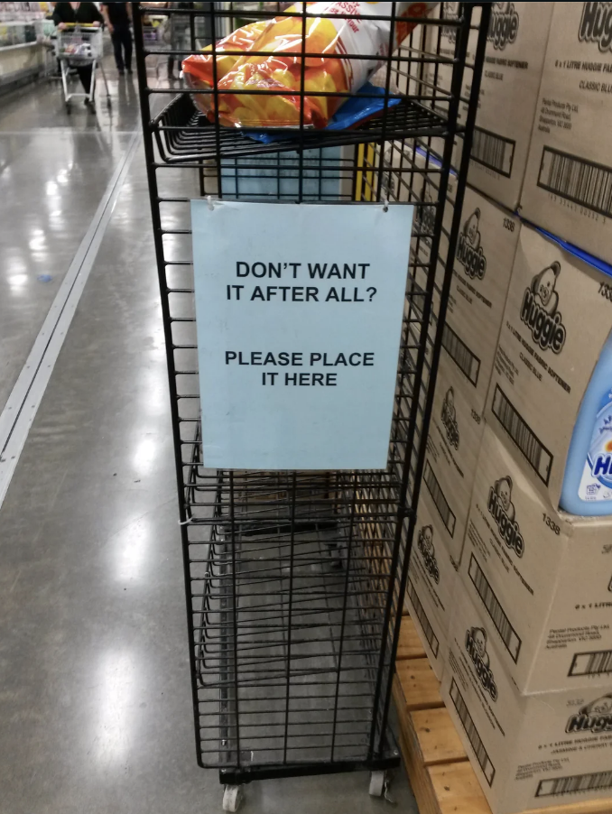 A sign in a store reads "DON'T WANT IT AFTER ALL? PLEASE PLACE IT HERE" on a metal rack with items returned by shoppers