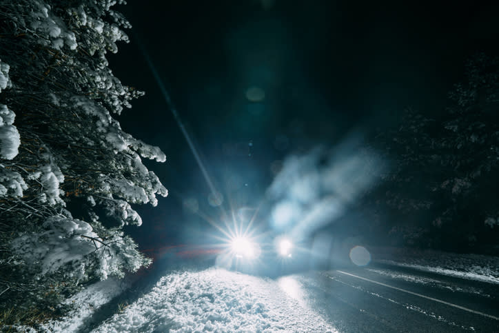 A car with bright headlights drives on a snowy road through a forest at night